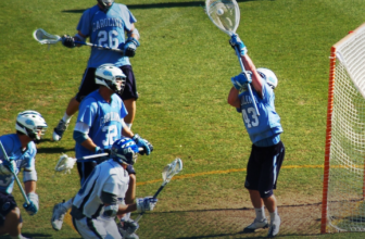 Lacrosse goalie in the crease raising the goalie stick to make a save, defenders in front of the net